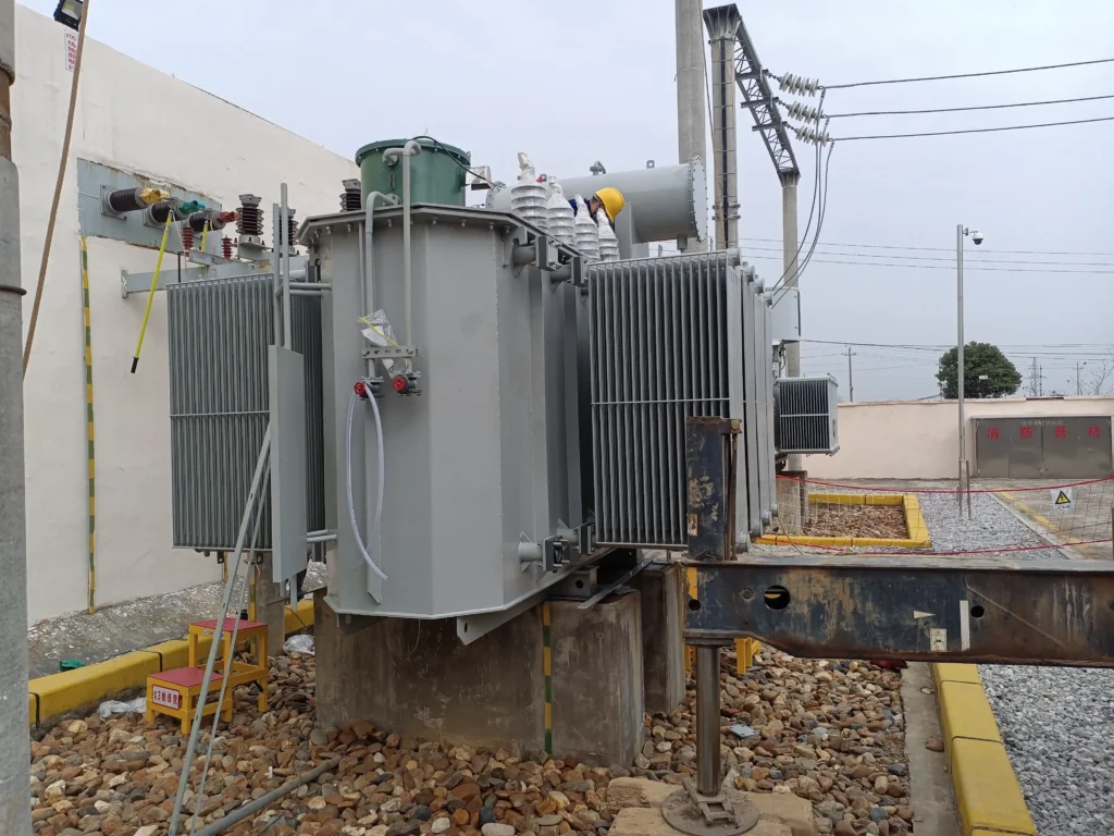 A field technician commissioning an oil immersed self cooled transformer at a substation, featuring corrugated radiator banks, concrete plinths, and a gravel containment pit by Baitong Electric.