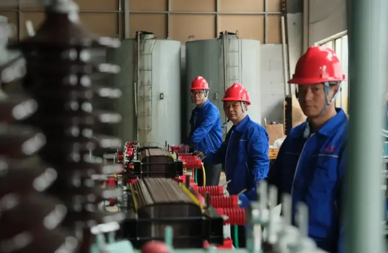 A dedicated transformer manufacturing team wearing blue uniforms and red hard hats assembling a core structure on the Baitong Electric factory floor.