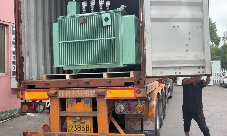 A factory worker carefully loading a large mint-green 22kv oil immersed transformer into an ocean shipping container on the back of an orange transport truck.